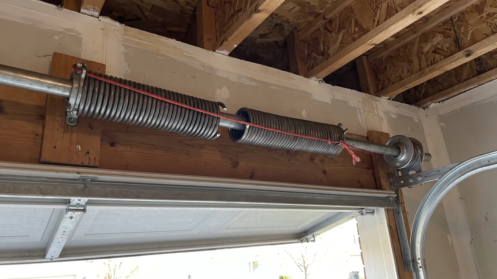Close-up of a snapped steel torsion spring on the shaft above a residential garage door, with a visible gap between the coils and the cable drum beside it