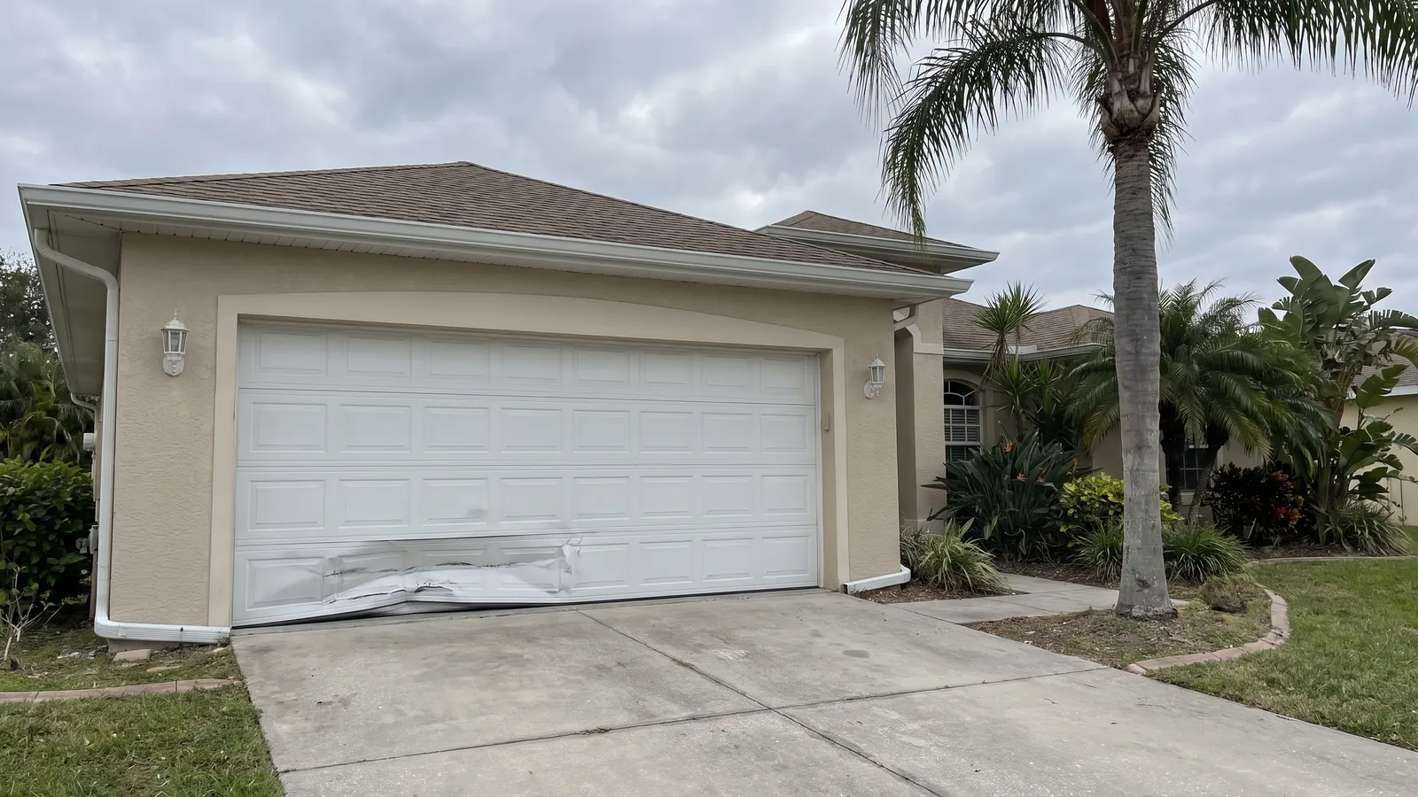 White steel garage door with the bottom panel dented and creased where a car backed into it, on a beige stucco Florida home
