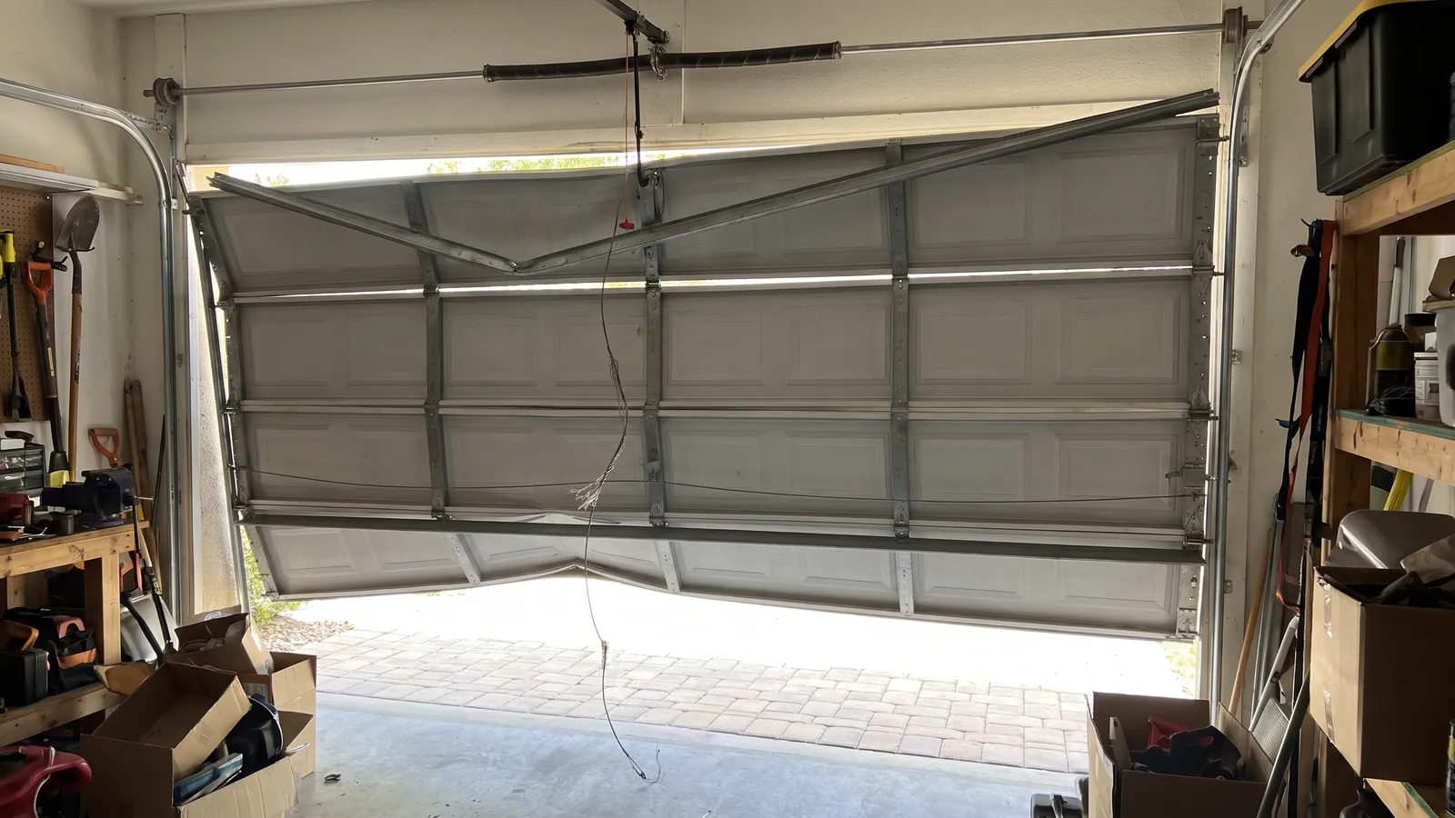 Sectional garage door hanging crooked and jammed off its track on one side with a frayed, snapped lift cable, seen from inside a Florida garage