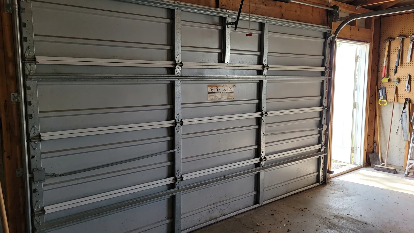 Inside view of a closed steel garage door showing horizontal reinforcing struts, heavy hinges, jamb brackets and the wind-load rating label
